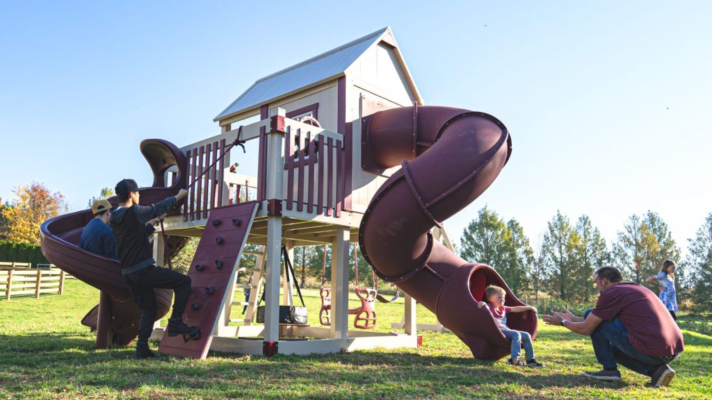 children playing on a large maroon and tan playset with swing set accessories including a climbing wall and two spiral tube slides